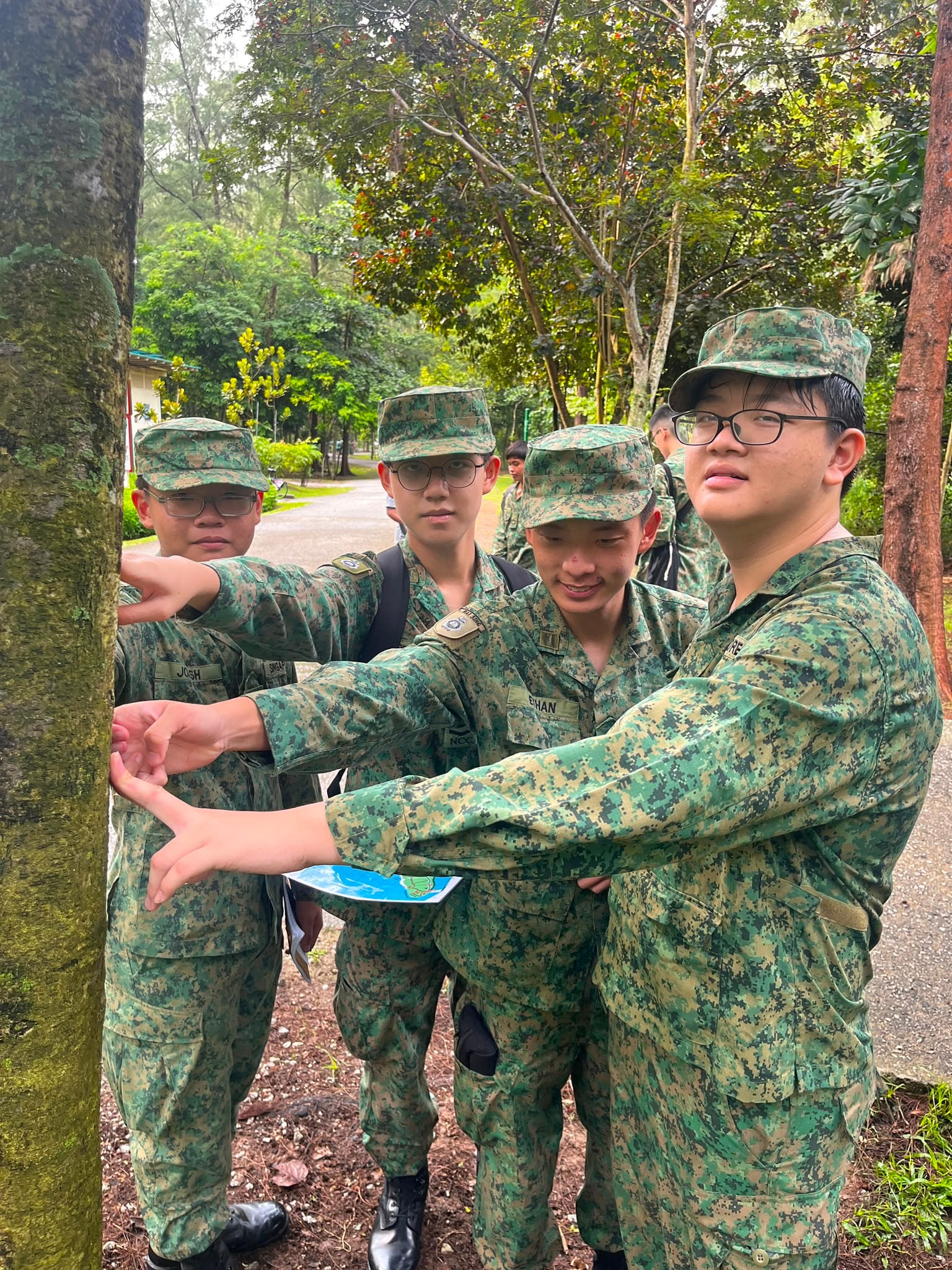 A group of NCC cadets in pixelated camouflage uniforms outdoor near trees, pointing at a tree trunk.